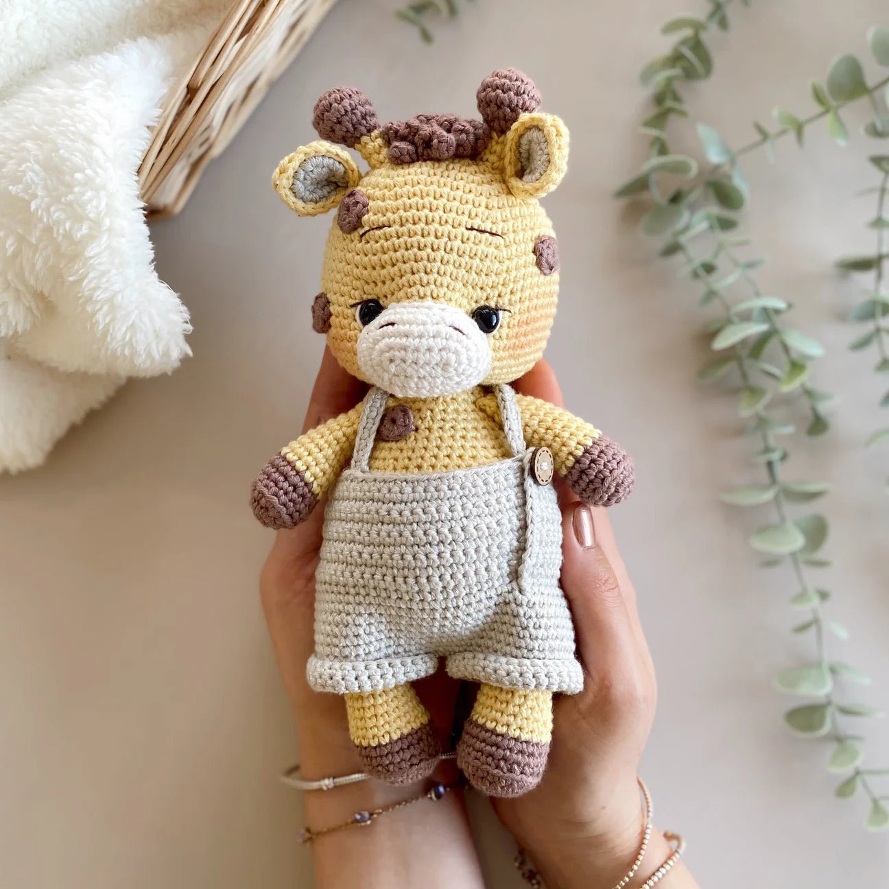 Crocheted yellow giraffe plushie wearing gray overalls, held by a hand with bracelets