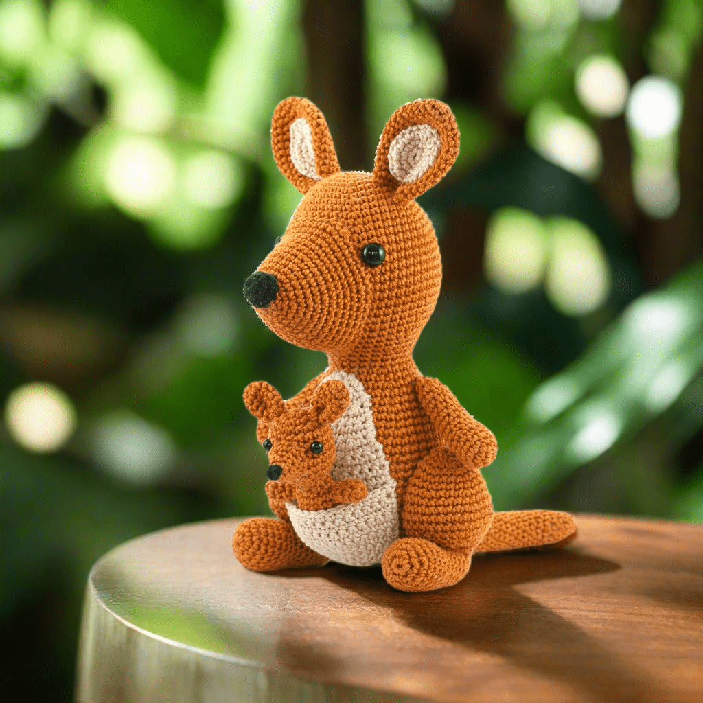 Crocheted orange kangaroo with baby joey sitting on wooden surface with blurred green background