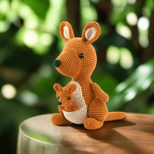 Crocheted orange kangaroo with baby joey sitting on wooden surface with blurred green background