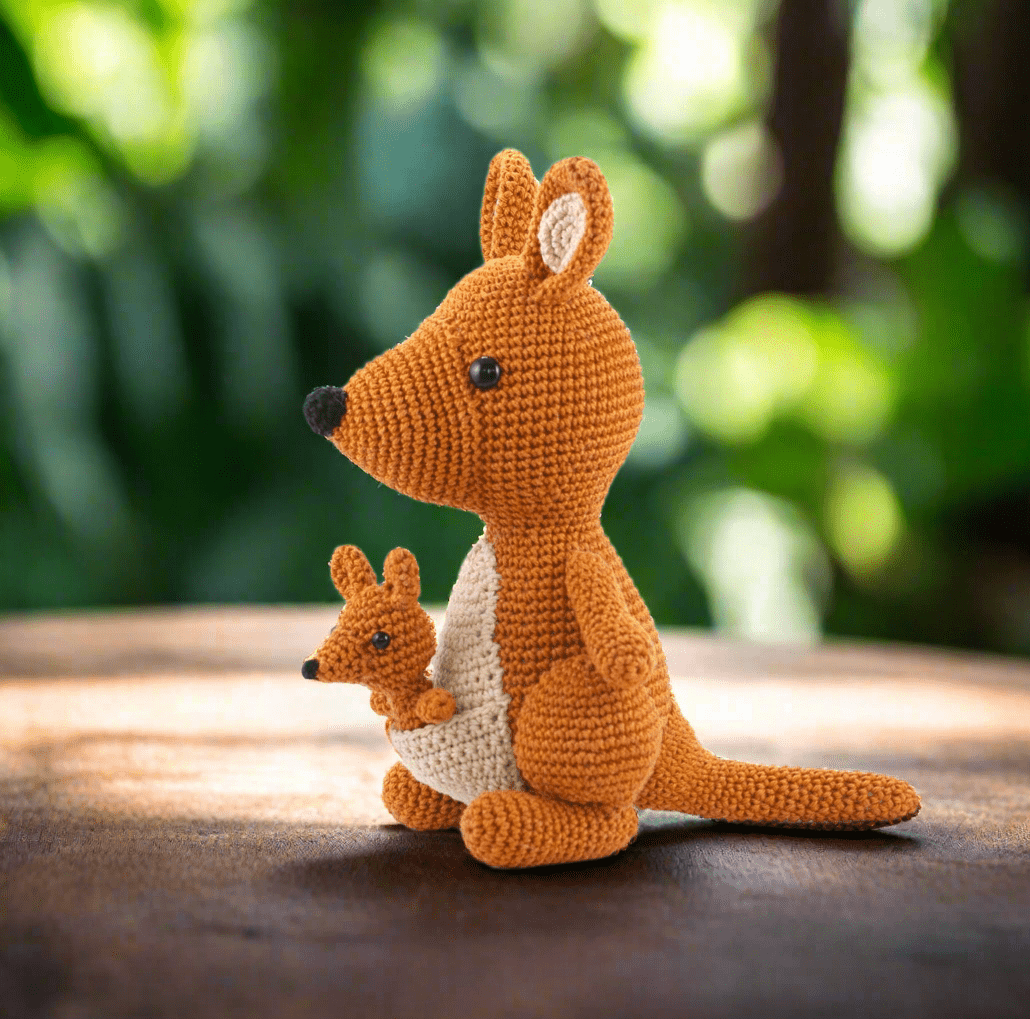 Crocheted orange kangaroo with baby joey, sitting on textured surface with blurred green background