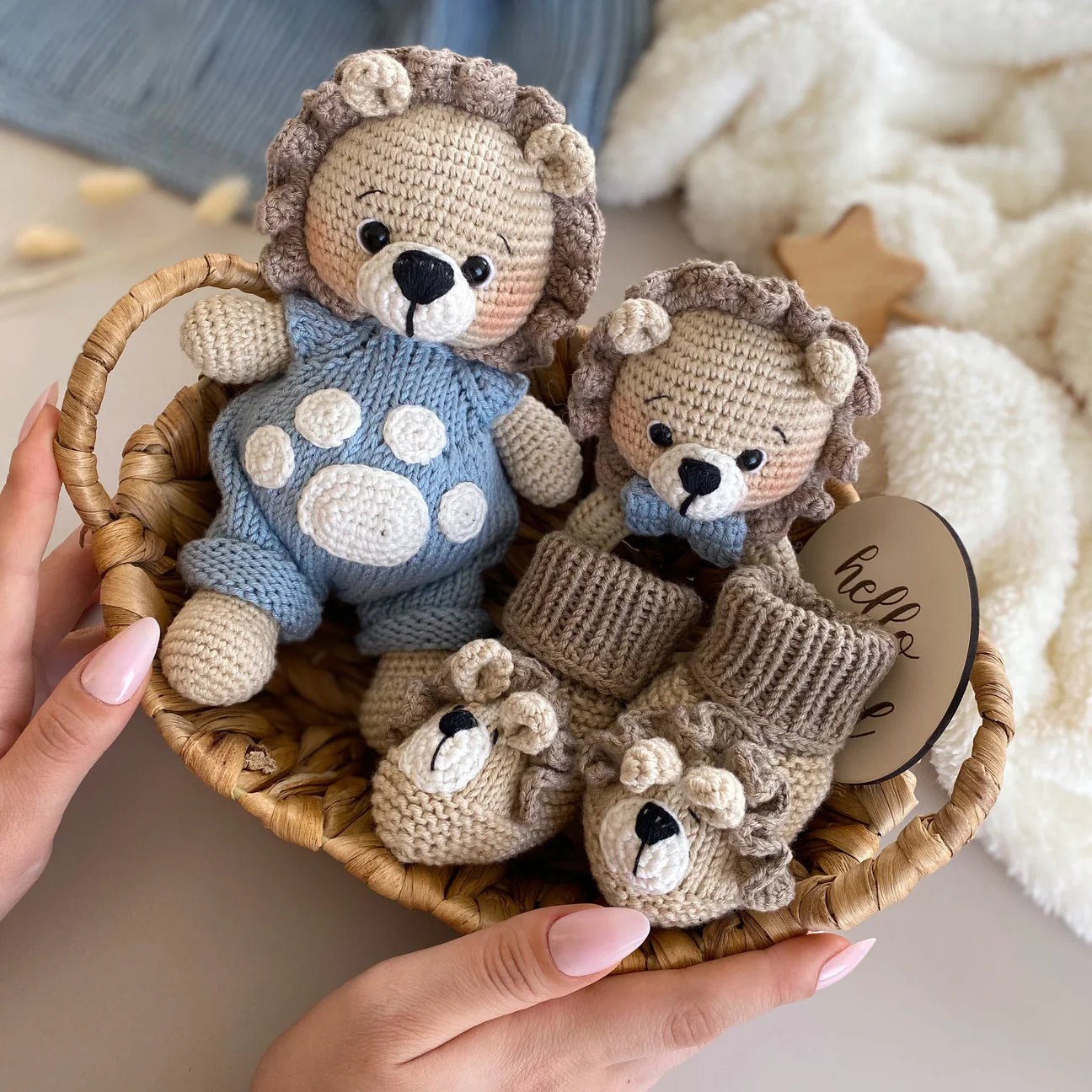 Crocheted lion stuffed animals in blue and beige, nestled in a woven basket with "hello" tag