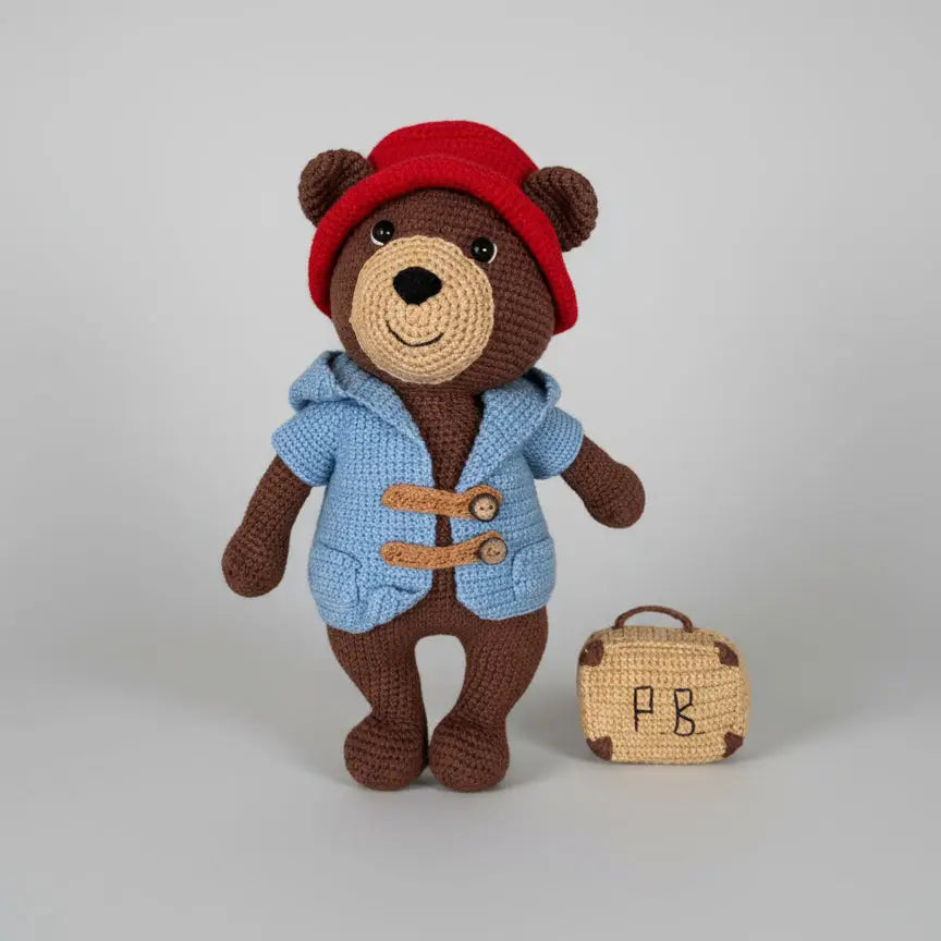 Crocheted teddy bear in blue duffle coat and red hat, standing next to a small matching suitcase with "PB" initials.