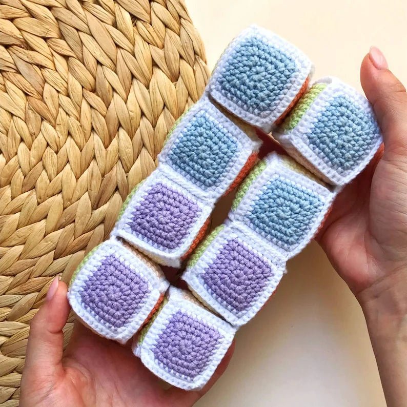 Crocheted square blocks in pastel blue and purple, held in hands over woven surface