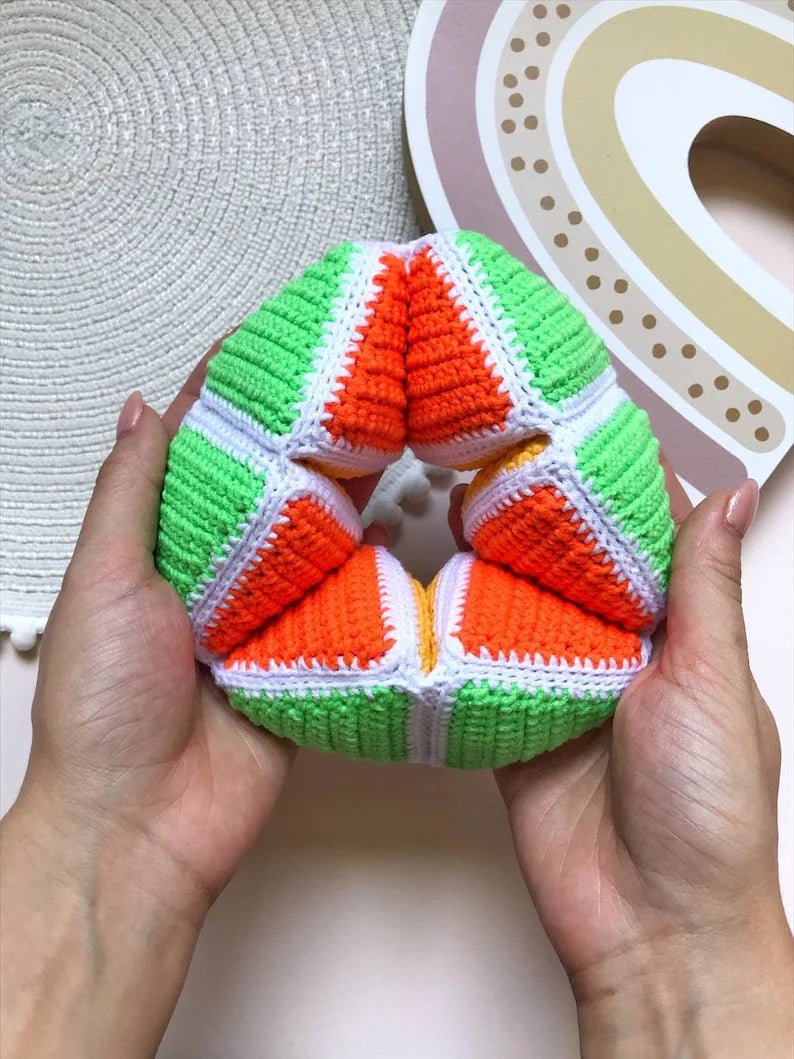 Crocheted watermelon-like geometric object in green, white, and orange, held between hands