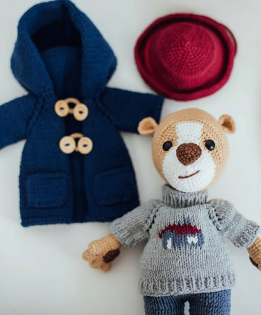 Knitted teddy bear in gray sweater next to blue coat and red hat