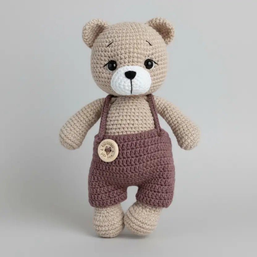 Crocheted teddy bear with brown overalls and a large button, standing against a neutral background