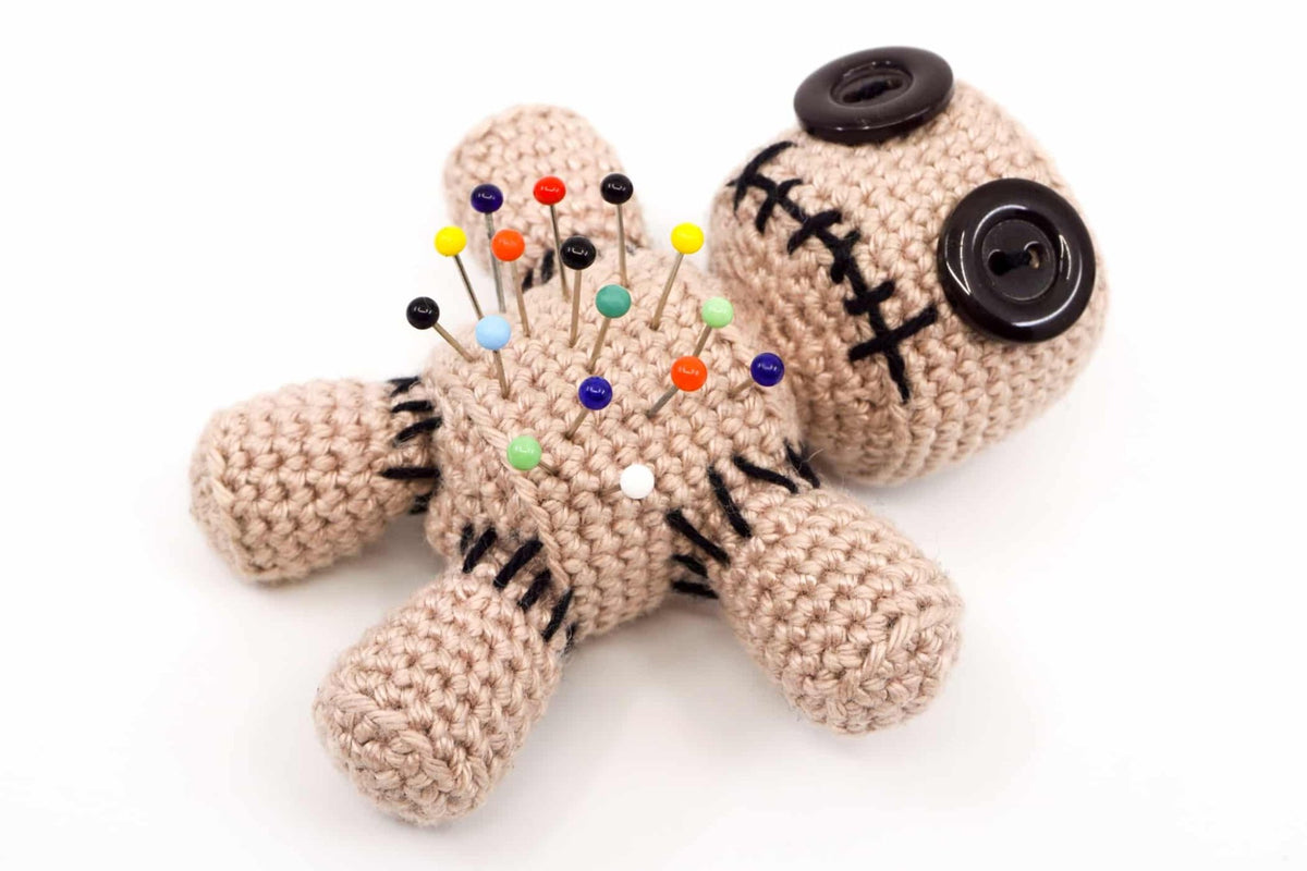 Crocheted voodoo doll with colorful pins and black buttons on beige yarn body