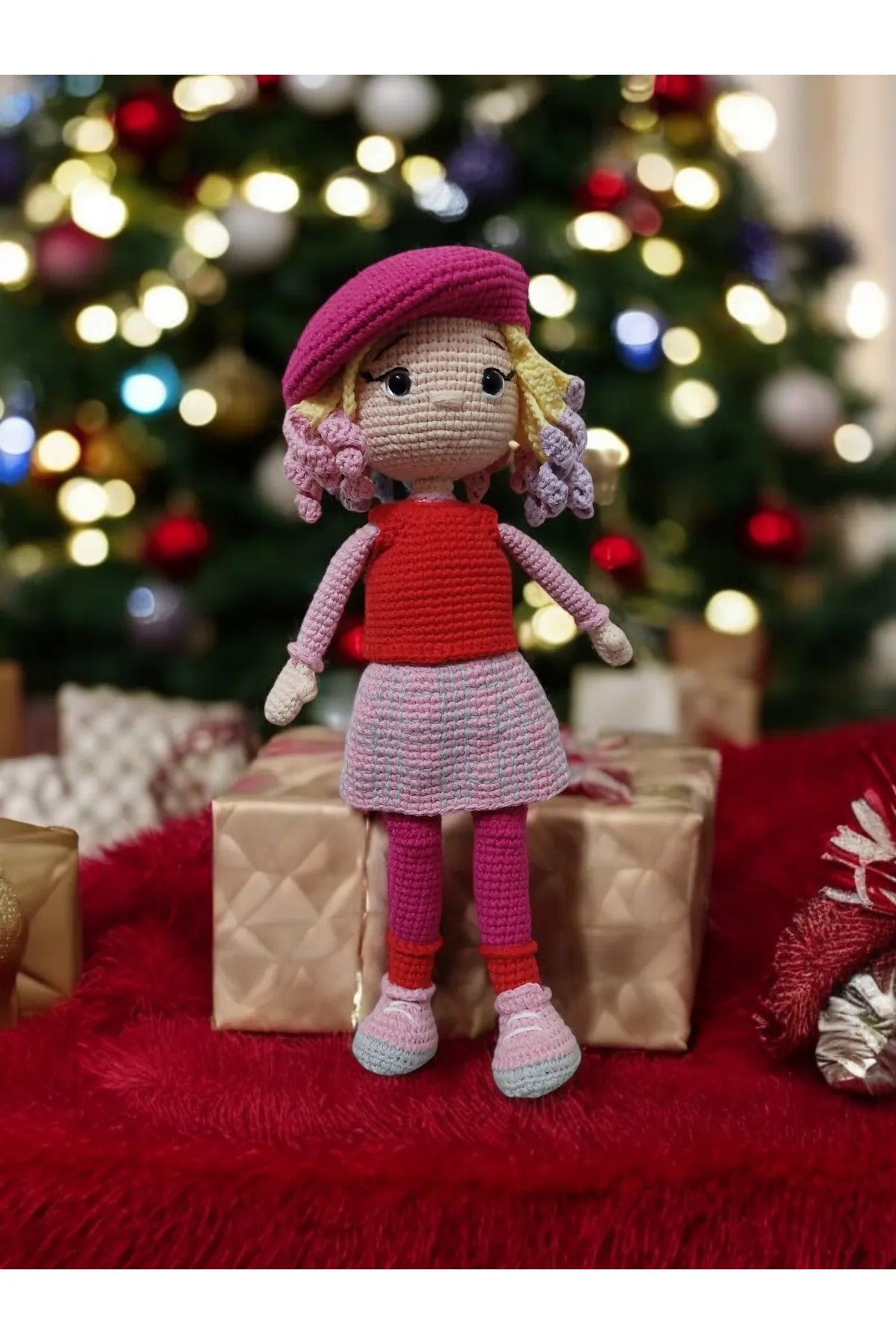 Crocheted doll with pink beret standing on red fur near wrapped Christmas gifts
