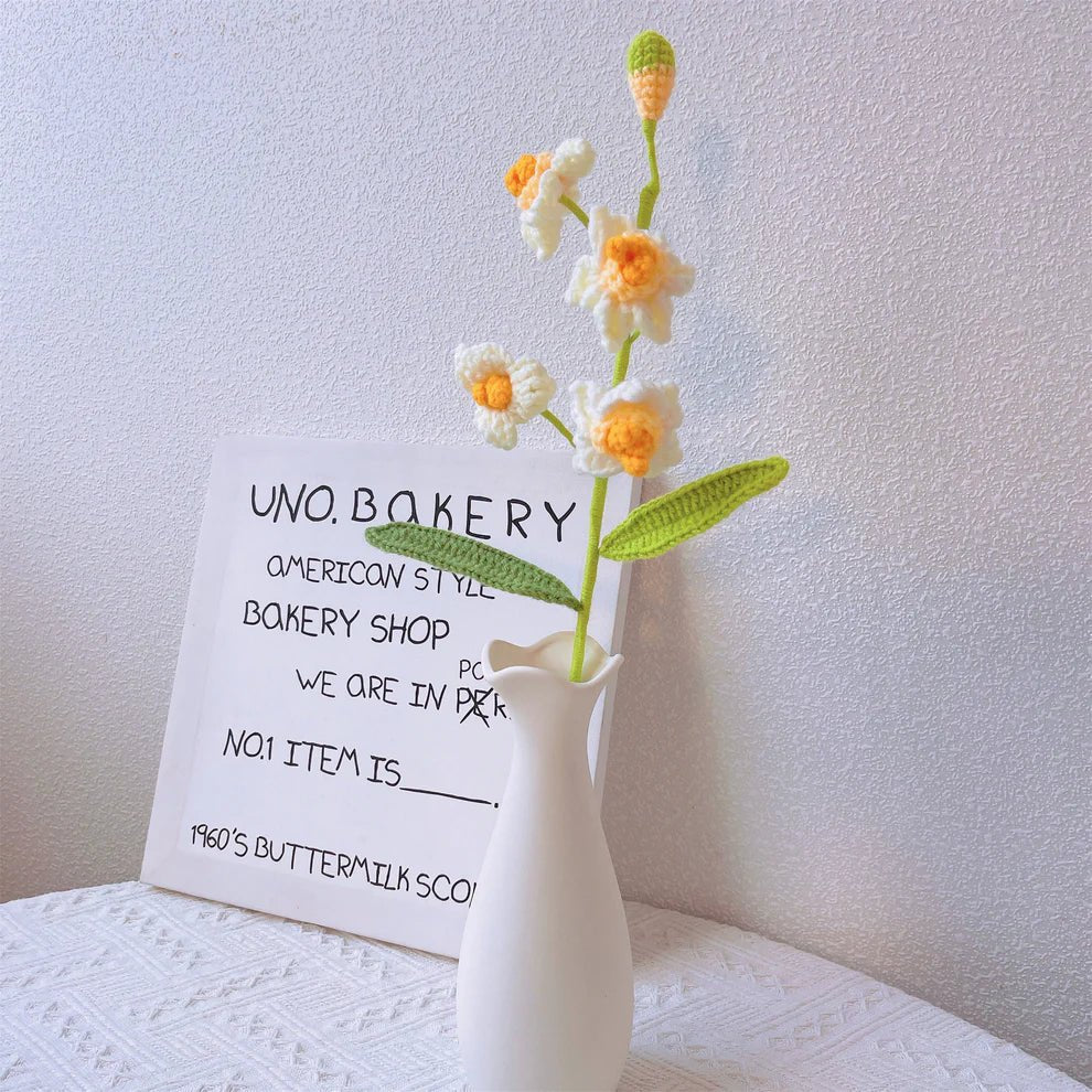 Crocheted daisies beside Uno Bakery sign in white ceramic vase on textured cloth
