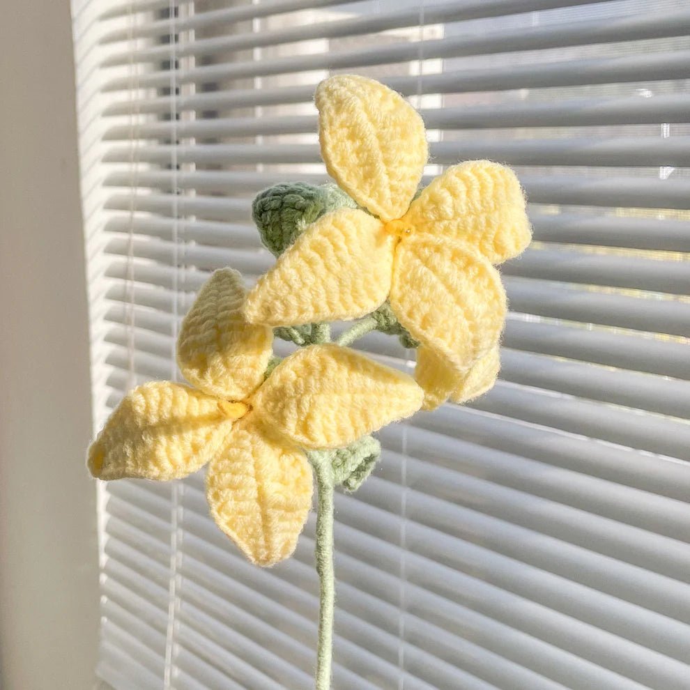 Crocheted yellow flower with green stem against white window blinds background