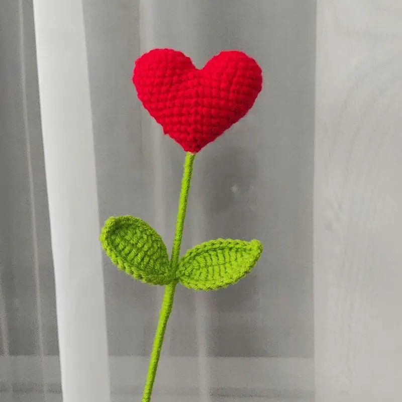 Crocheted red heart-shaped flower with green stem and leaves on gray background