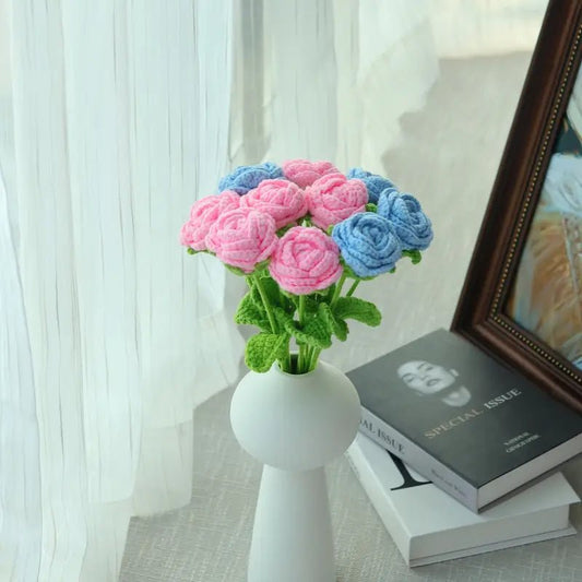 Crocheted pink and blue roses in white vase beside books near curtained window