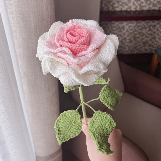 Delicate crocheted rose in white and pink, with green leaves, held against curtain