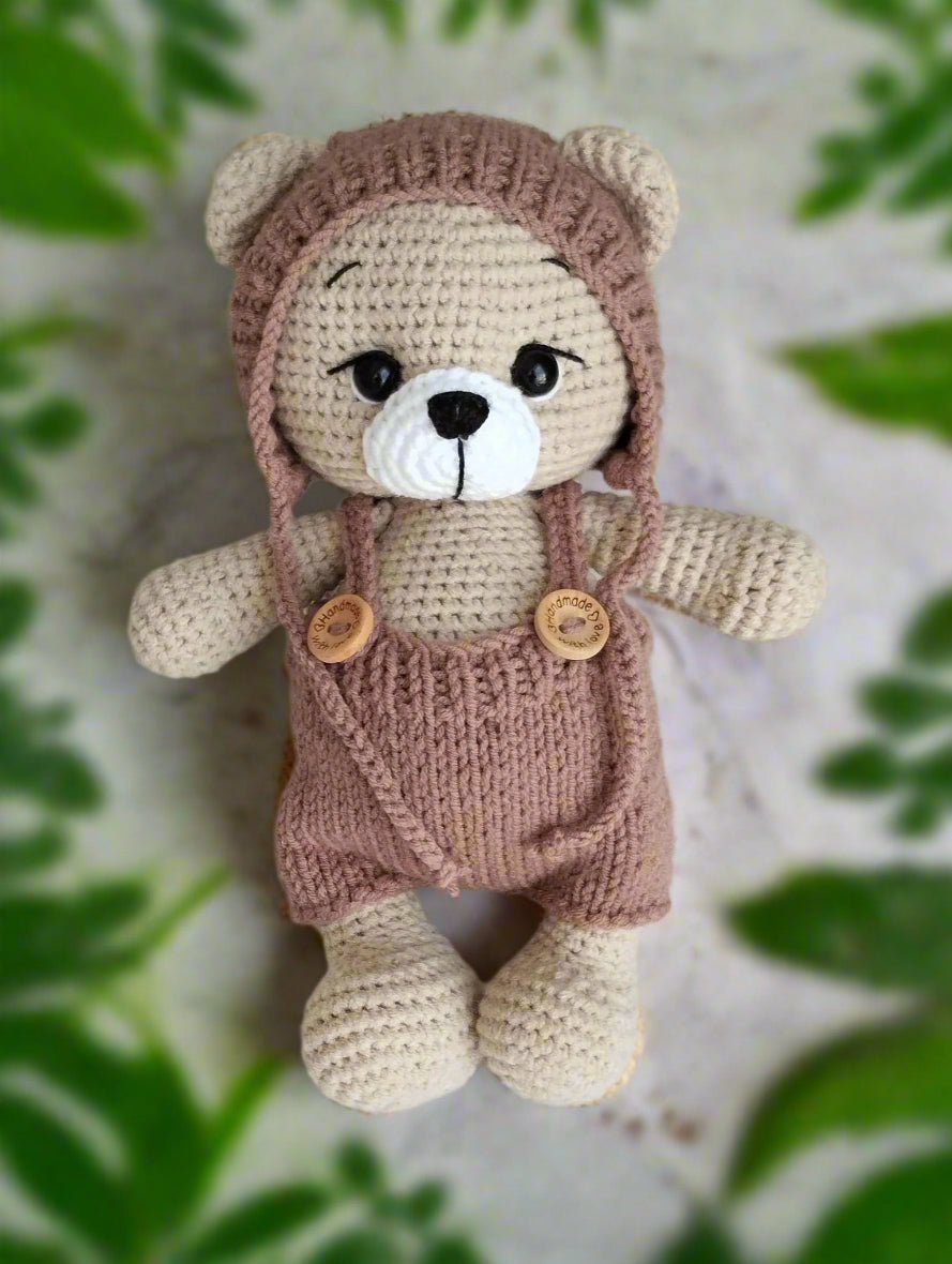 Crocheted teddy bear in dusty pink overalls with wooden buttons, soft beige color