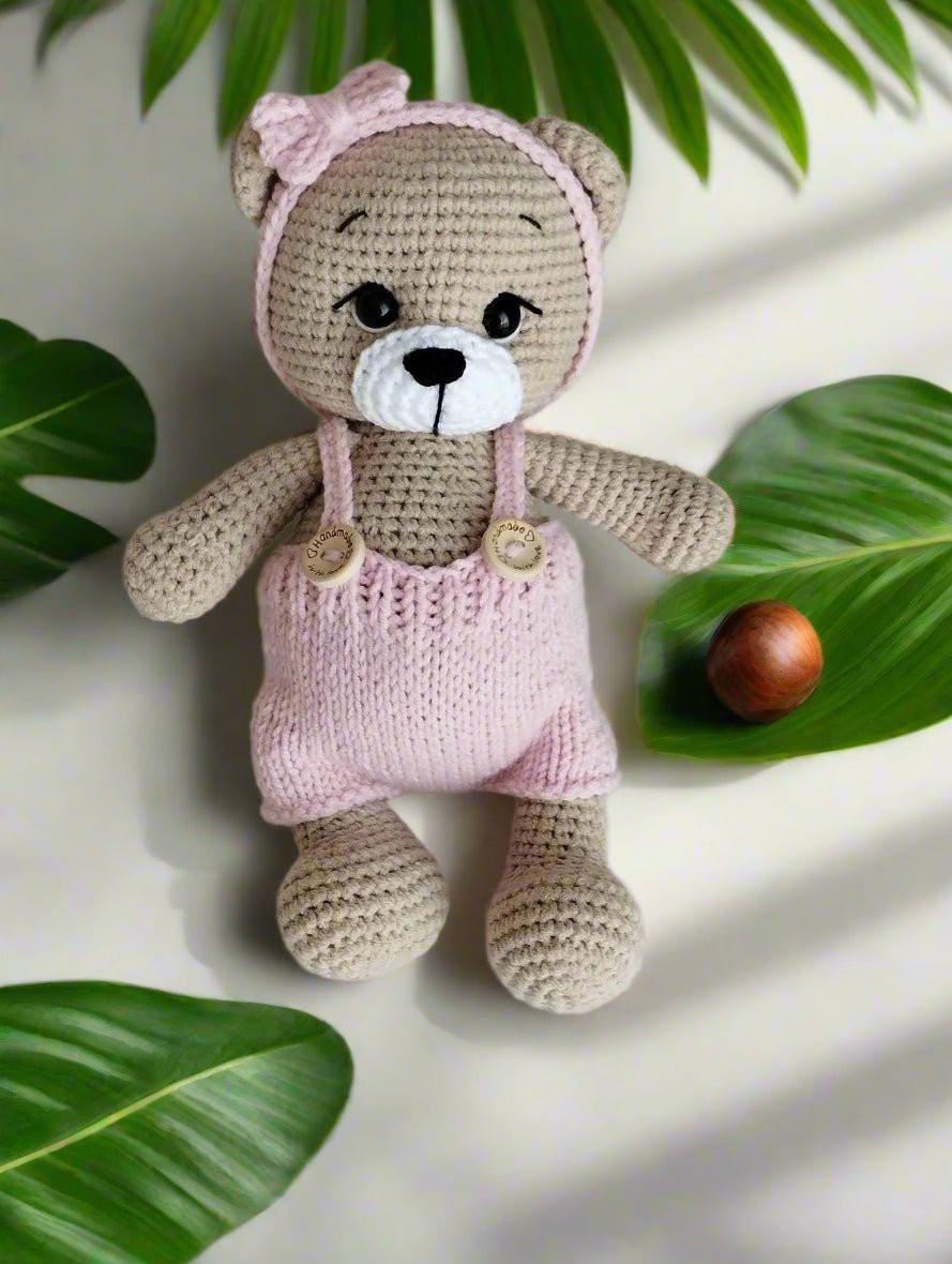 Handmade crocheted teddy bear in soft pink and beige, sitting among green leaves with a wooden bead nearby.