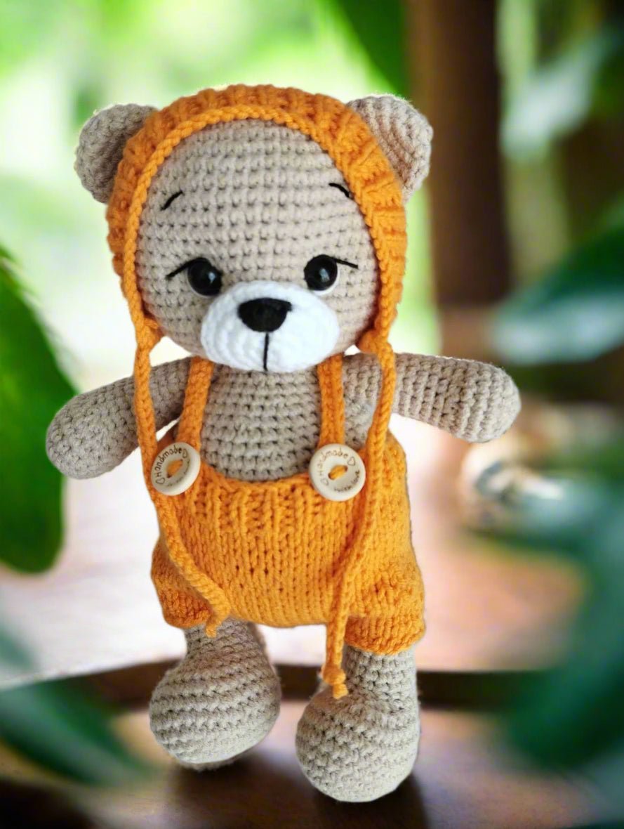 Crocheted teddy bear in bright orange overalls, gray body, cute button nose, sitting against blurred green background.