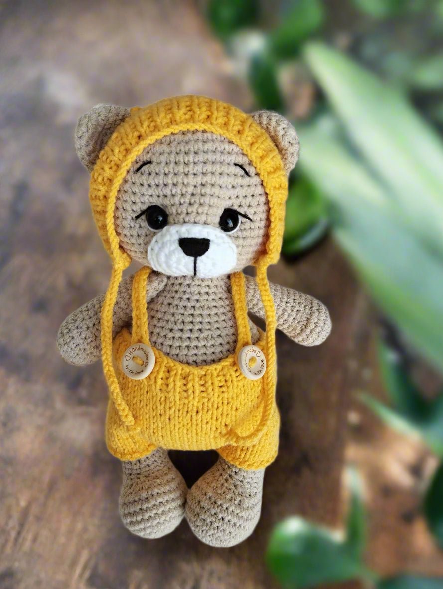 Crocheted teddy bear in yellow overalls with cute black button eyes, sitting on wooden surface with blurred green background