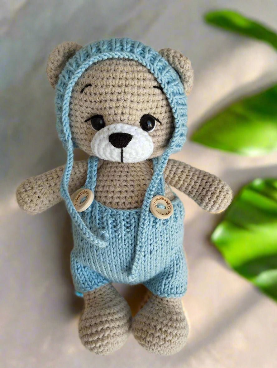 Crocheted teddy bear in blue knitted overalls and hat, with cute button details and soft gray yarn.