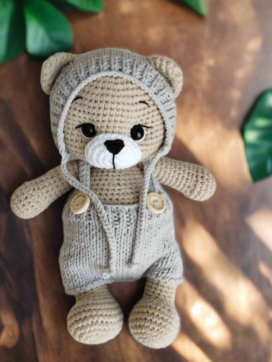Crocheted teddy bear in soft gray yarn, wearing a knitted hoodie and overalls with wooden buttons.