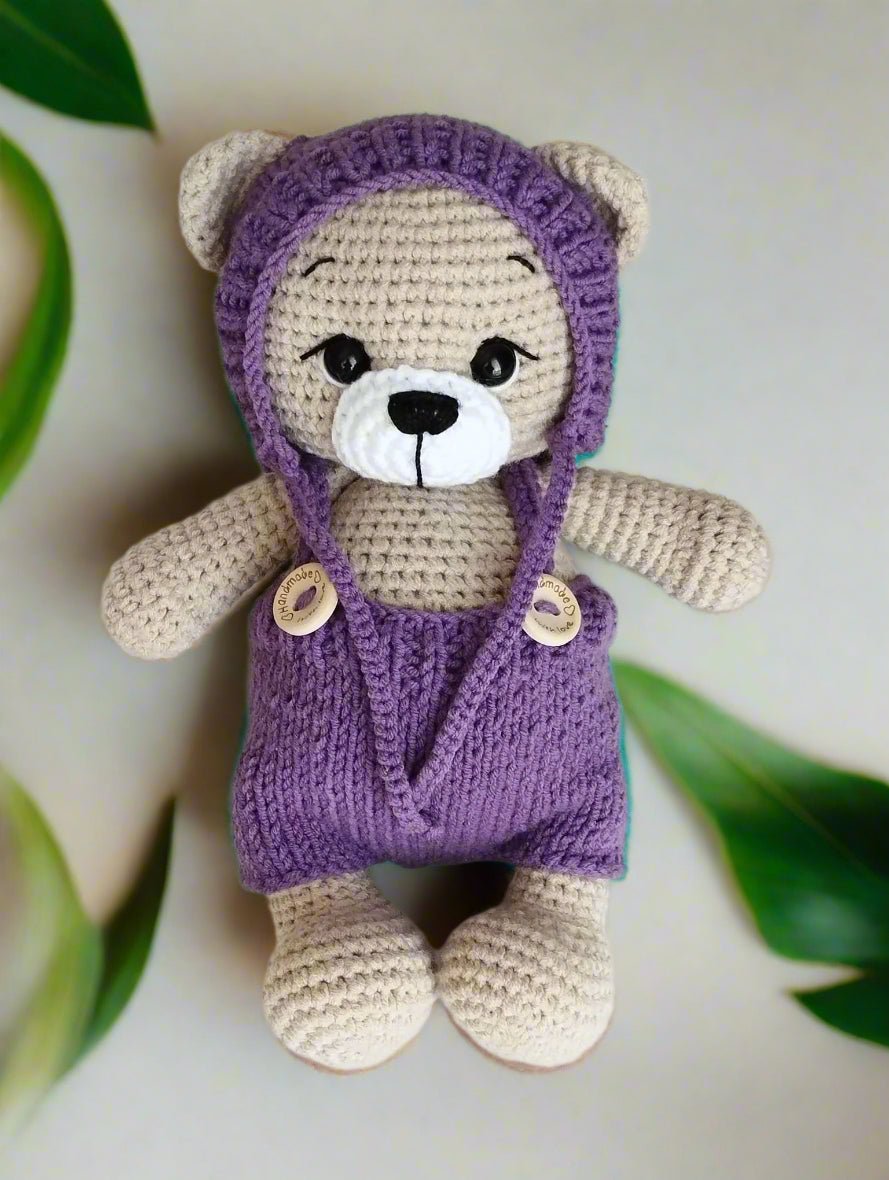 Crocheted teddy bear in purple overalls with wooden buttons and green leaves