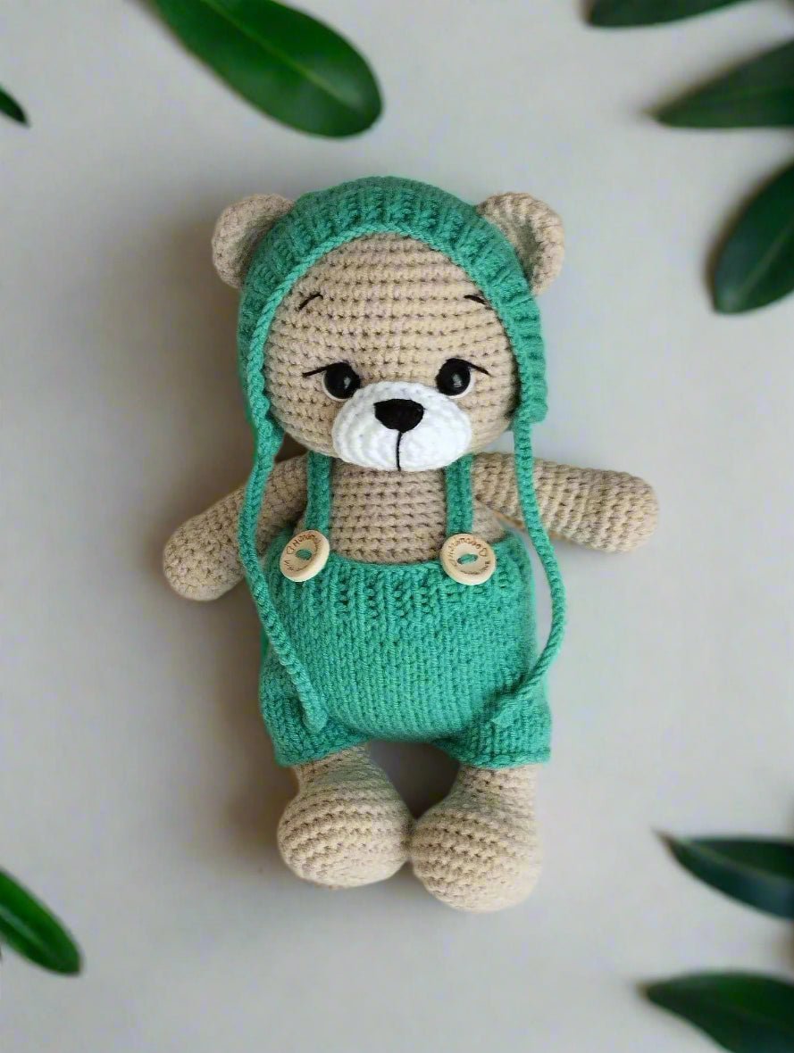 Crocheted teddy bear in mint green overalls and hood, with beige body and cute button nose, sitting against a soft background.
