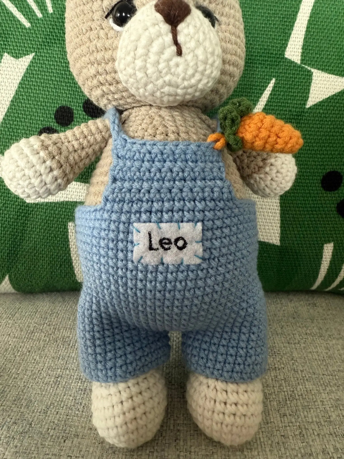 Crocheted teddy bear in blue overalls, holding an orange carrot, named Leo