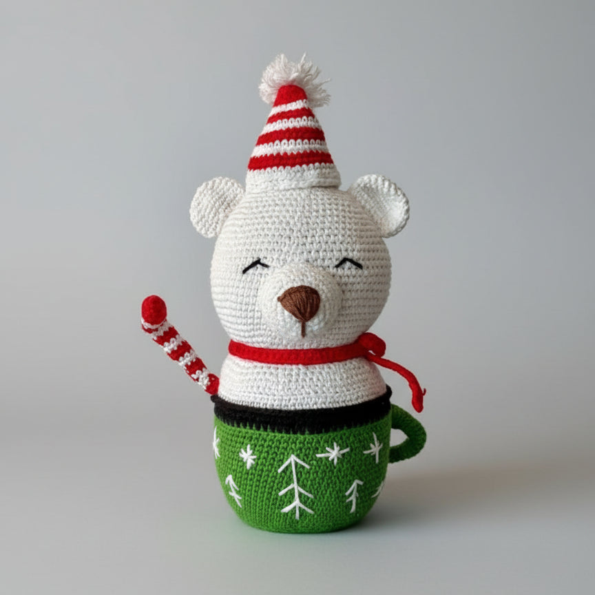 Crocheted white teddy bear with red striped hat and scarf, sitting in a green Christmas-themed mug with snowflake pattern