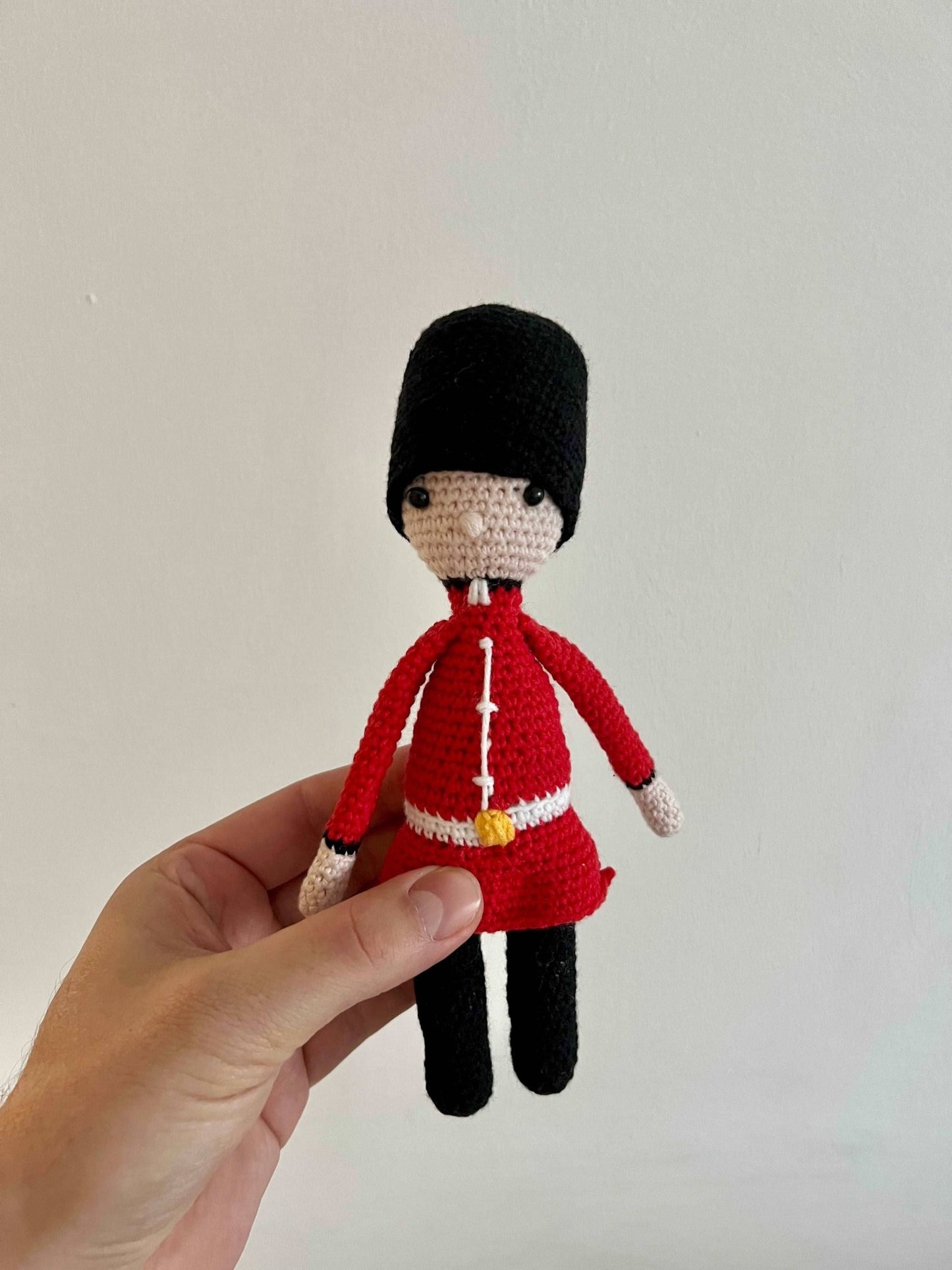 Crocheted Royal Guard figurine in red uniform and black bearskin hat, held by a hand against a light background