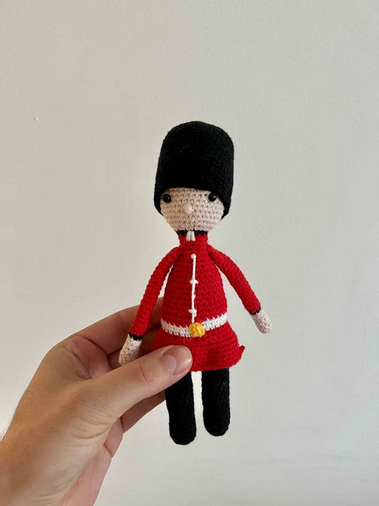 Crocheted Royal Guard figurine in red uniform and black bearskin hat, held by a hand against a light background