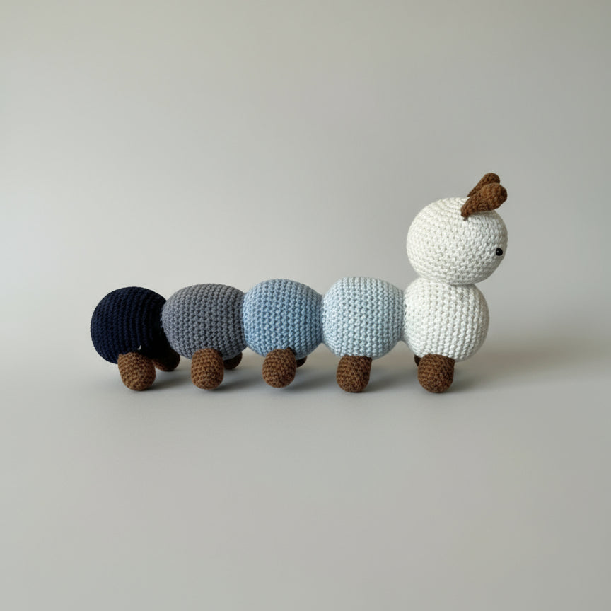 Crocheted caterpillar-like creature in navy, gray, light blue, and white, with brown feet, against neutral background