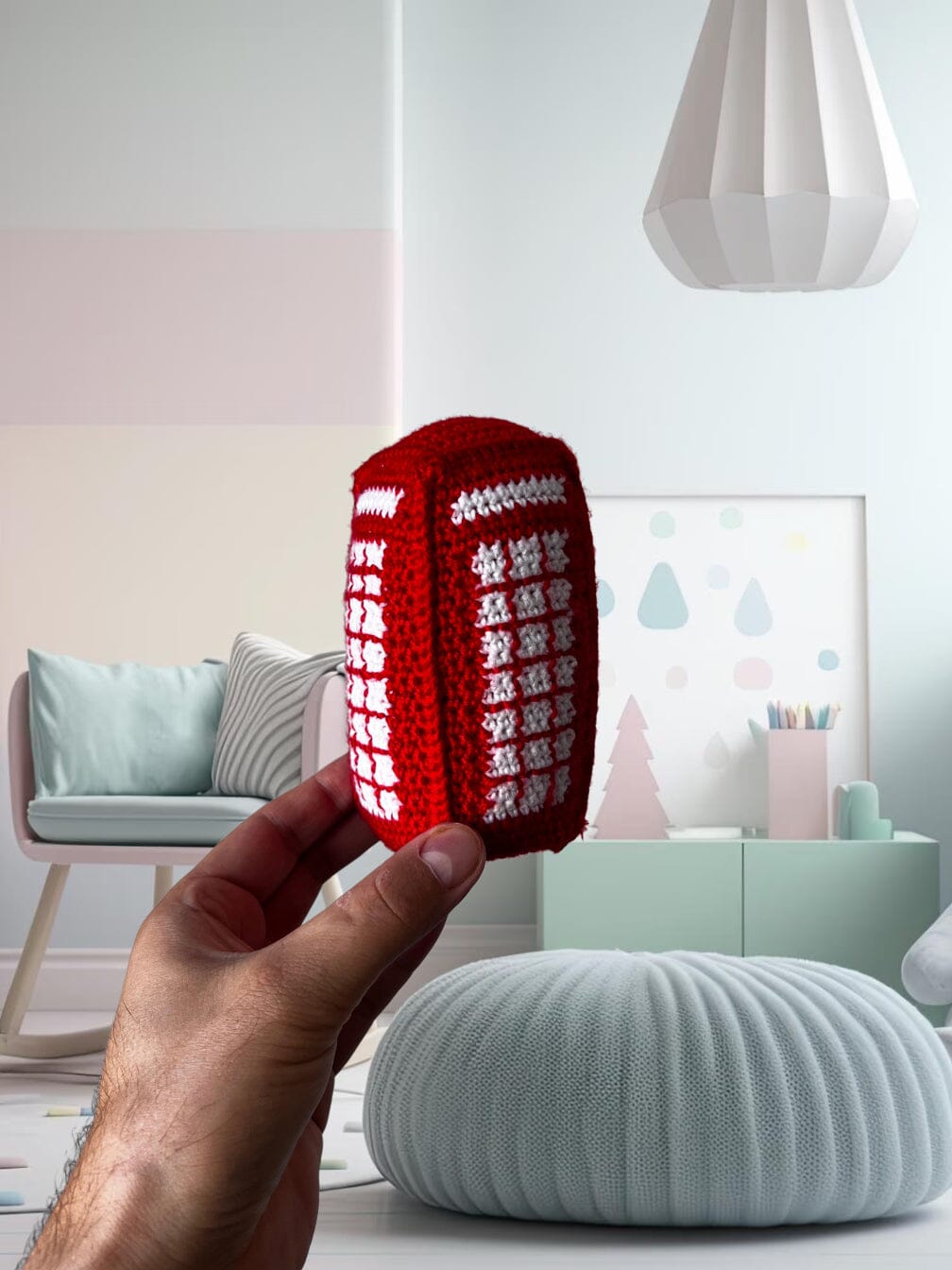 Hand holding crocheted red telephone booth with white details, against a pastel minimalist interior background with soft furniture and decor.