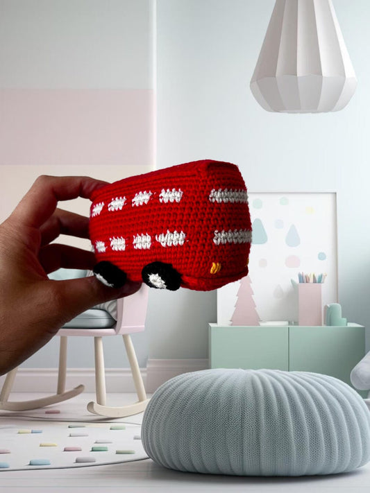 Hand holding a red crocheted bus with white and black details in a minimalist, pastel-colored nursery setting.
