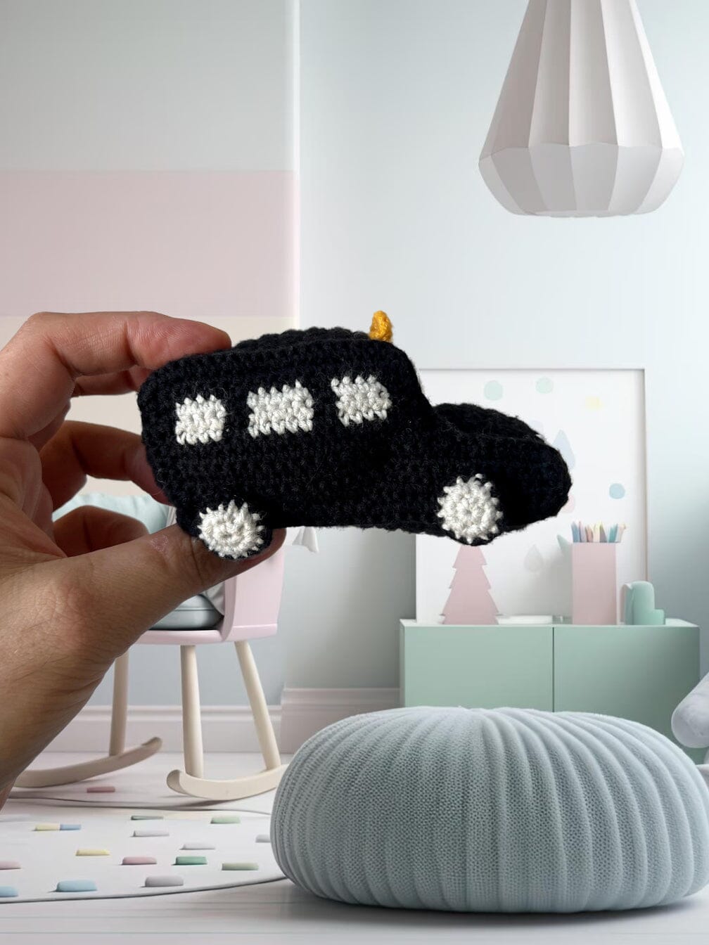 Hand holding a black crocheted car with white windows and headlights against a pastel minimalist nursery background.