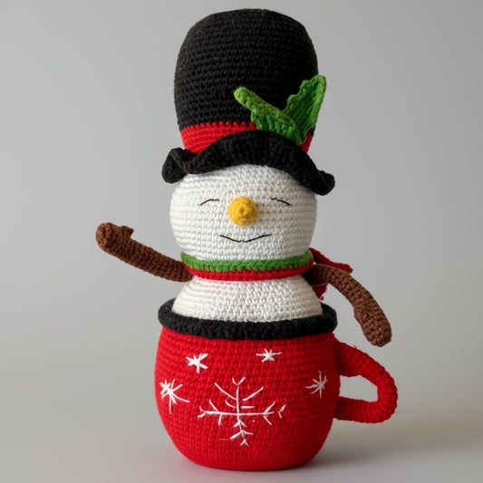 Crocheted snowman with black top hat, nestled in a red mug with snowflake design, wearing a green and red scarf.